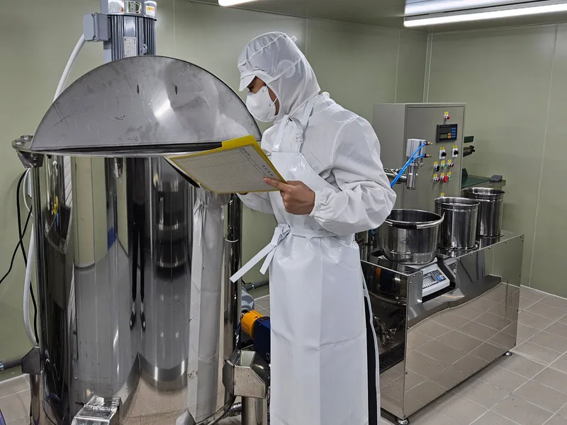 Worker in full sanitary gear checking a batch log beside a stainless mixing tank at the ANGHODU plant
